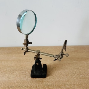 May include: A metal helping hand tool featuring a circular magnifying glass with a silver frame, mounted on an adjustable arm with multiple wing nuts for positioning. The device includes a single alligator clip and a heavy black rectangular base.