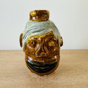 May include: A ceramic jug shaped like a human head with a glossy brown glaze. The figure features a wide black mouth, a prominent nose, and a textured light blue-grey glaze forming hair across the forehead. The vessel has a cylindrical neck opening.