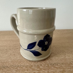 May include: A beige ceramic mug with a dark blue floral design. The mug has a cylindrical shape with a handle and a decorative band around the middle. The floral design features a flower and leaves.