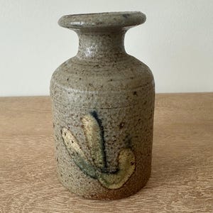 May include: A handcrafted, stoneware vase with a textured, speckled surface. The vase is cylindrical with a flared rim and a decorative, abstract design in shades of green and blue. The vase is approximately 6 inches tall.