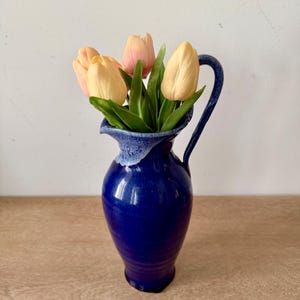 May include: A deep blue ceramic pitcher with a glossy finish and a lighter blue drip glaze around the rim. The pitcher features a curved handle and a flared spout, and it is filled with a bouquet of artificial peach and yellow tulip flowers.