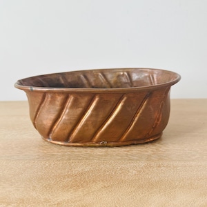 May include: An oval-shaped copper bowl featuring a series of diagonal, raised ridges along its exterior walls. The metal has a warm, metallic brown finish with visible variations in tone and texture across the curved surface of the vessel.