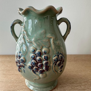 May include: A ceramic vase in a light green hue, adorned with embossed grape clusters and leaves. The vase features two handles and a ruffled rim. The grapes are a deep blue color. The vase is a decorative item.
