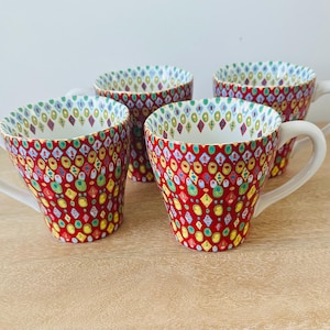 May include: A set of four ceramic mugs featuring a vibrant red base color with a repeating pattern of multicolored geometric shapes including diamonds and ovals in shades of yellow, green, and blue. Each mug has a white interior and a white handle.