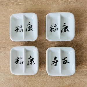 May include: Four white ceramic square sauce dishes, each featuring a central divider. Three dishes display the black Chinese characters 福康, while the fourth dish features the black Chinese characters 寿友. The dishes have rounded corners.