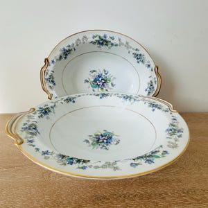 May include: Two white porcelain serving bowls with gold-trimmed edges and handles. The bowls feature a delicate floral pattern of blue and purple flowers with green leaves, centered in the basin and repeated along the wide, curved rims.