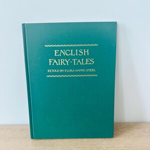 May include: A teal green hardcover book with the title "ENGLISH FAIRY TALES" in gold lettering. The author's name, "RETOLD BY FLORA ANNIE STEEL," is also in gold. The book is resting on a light-colored wooden surface.