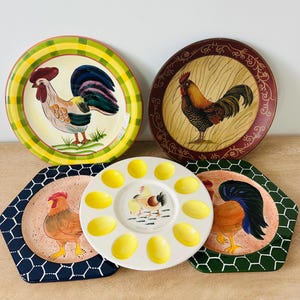 May include: A collection of five ceramic plates featuring rooster motifs. Two round plates have yellow plaid and dark red borders. Two hexagonal plates have dark blue and green chicken wire patterns. One round white egg serving plate is centered.
