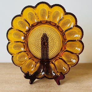 May include: An amber-colored glass egg plate featuring a central raised pillar and a circular arrangement of twelve individual egg-shaped indentations. The surface is decorated with a textured hobnail pattern throughout the entire plate.