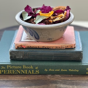 May include: A small, gray stoneware bowl filled with dried flowers and herbs, resting atop a stack of vintage books. The bowl has a blue floral design and is marked with "Pottery Works" and the number "1932". The books are various colors and sizes.