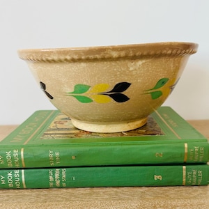 May include: A tan ceramic bowl with a decorative leaf pattern in green, yellow, and black, resting on two green books. The bowl has a textured rim and a light-colored base. The books are titled "My Book House".