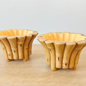 May include: Two round planters featuring a textured exterior designed to resemble vertical bamboo stalks in shades of tan and brown. The interior of each planter is a smooth, pale yellow color, and the base is supported by small, integrated feet.