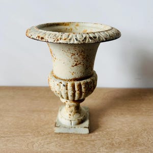 May include: An off-white cast iron urn with a wide, flared rim featuring decorative detailing. The urn has a bulbous, ribbed body and a square base, with visible patches of brown rust throughout the surface, giving it a weathered appearance.