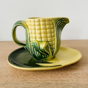 May include: A ceramic creamer pitcher and matching saucer shaped like an ear of corn. The pitcher features a yellow kernel body with green husk details and a curved handle. The saucer is shaped like a corn leaf with a yellow and green finish.