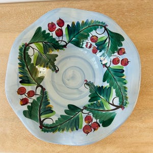 May include: A light blue ceramic serving bowl with a hand-painted design. The bowl features a wreath of green leaves and red berries around the rim. The interior has a light blue swirl pattern.