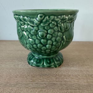 May include: A glossy, dark green ceramic planter featuring a raised, textured relief pattern of grape clusters and leaves around the exterior. The vessel has a wide, flared rim and sits on a circular, scalloped pedestal base with a crackle glaze finish.