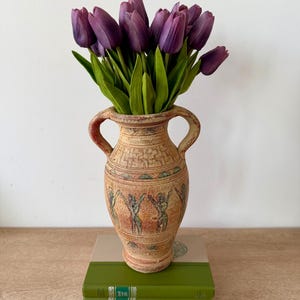 May include: A beige ceramic amphora vase with two handles, featuring a textured surface with geometric patterns and a frieze of stylized figures. The vase is filled with a bouquet of deep purple tulips with green stems and leaves.