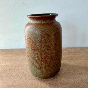May include: A cylindrical ceramic vase with a speckled brown and olive green finish. The surface features an embossed leaf pattern that wraps around the body. The vase has a wide, rounded opening with a dark interior rim and a smooth, curved neck.