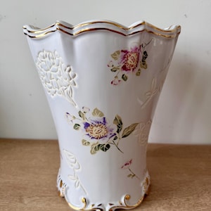Puede incluir: Un jarrón de cerámica blanca con borde y base festoneados, ambos con detalles en oro. La superficie presenta patrones florales en relieve y motivos de flores pintadas en tonos morado, rosa y verde, creando una apariencia texturizada.