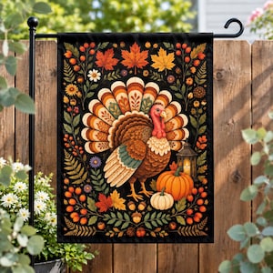 May include: A rectangular garden flag featuring a central turkey with a fanned tail of orange, brown, and cream feathers. The design includes autumn maple leaves, berries, acorns, small flowers, two pumpkins, and a lit lantern on a black background.
