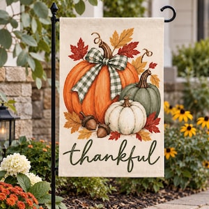 May include: A rectangular garden flag featuring a large orange pumpkin with a green and white checkered bow, a smaller white pumpkin, and a sage green pumpkin. Two brown acorns and autumn leaves surround the pumpkins, with the word thankful in script.