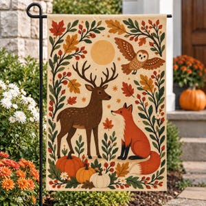 May include: A rectangular garden flag featuring a cream background with a central illustration of a brown deer and an orange fox. The scene includes a flying owl, a yellow sun, pumpkins, autumn leaves, and berries framed by green foliage.