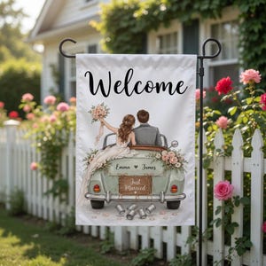 May include: A white garden flag with the word "Welcome" in black script. The flag features an illustration of a just-married couple in a vintage car, with the names "Emma" and "James" and the words "Just Married". The car is decorated with flowers.