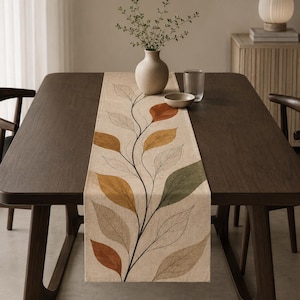 May include: A rectangular beige table runner featuring a central black vine pattern with alternating leaves in shades of burnt orange, mustard yellow, sage green, and light gray, laid flat across the center of a dark brown wooden dining table.