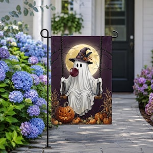 May include: A rectangular garden flag featuring a white ghost wearing a witch hat and blowing a bubble, sitting on a swing against a full moon background. The scene includes a large jack-o'-lantern and smaller pumpkins at the base.