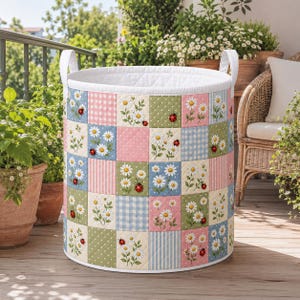 May include: A cylindrical quilted fabric storage basket featuring a patchwork design of alternating squares. The pattern includes white daisies, red ladybugs, and various pastel backgrounds in pink, blue, and green with gingham and polka dot prints.