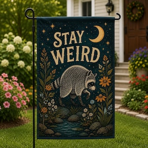 May include: A dark blue garden flag featuring a raccoon walking through a floral landscape with rocks and a stream. The text STAY WEIRD is printed in large cream letters above the raccoon, accompanied by a yellow crescent moon and stars.