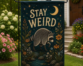 May include: A dark blue garden flag featuring a raccoon walking through a floral landscape with rocks and a stream. The text STAY WEIRD is printed in large cream letters above the raccoon, accompanied by a yellow crescent moon and stars.