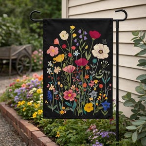 May include: A black garden flag featuring a colorful floral design. The flowers include pink, yellow, red, and white blooms, with green stems and leaves. A small butterfly is also included in the design. The flag is displayed on a black metal stand.
