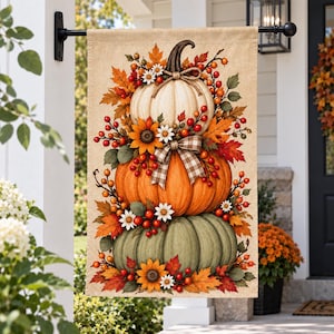 May include: A rectangular garden flag featuring a stack of three pumpkins in white, orange, and sage green. The design is decorated with autumn leaves, red berries, white daisies, and yellow sunflowers, accented by a brown and white gingham ribbon.