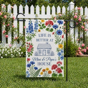 May include: A garden flag with the text "LIFE IS BETTER AT Mini & Papa's HOUSE" surrounded by colorful floral illustrations. The flag is displayed on a black metal stand in a garden setting with a white picket fence and blooming flowers in the background.