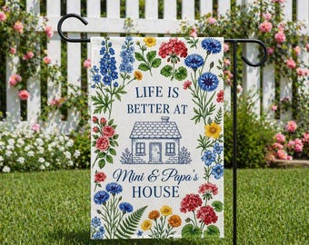 May include: A garden flag with the text "LIFE IS BETTER AT Mini & Papa's HOUSE" surrounded by colorful floral illustrations. The flag is displayed on a black metal stand in a garden setting with a white picket fence and blooming flowers in the background.