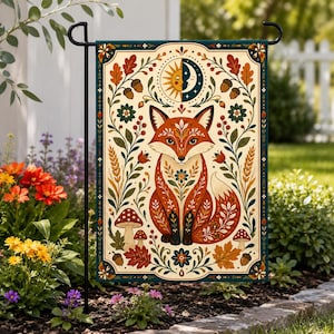 May include: A rectangular garden flag featuring a central illustration of a fox surrounded by botanical elements, mushrooms, acorns, and a sun and moon motif. The design uses a cream background with orange, red, green, and dark blue accents.
