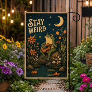 May include: A garden flag featuring a whimsical illustration of a frog playing a guitar. The flag has the words "STAY WEIRD" in a vintage font. The frog is sitting on a rock in a pond with flowers and a crescent moon in the background.