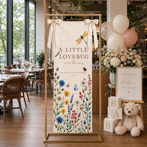 May include: A vertical cream-colored fabric banner suspended from a gold metal frame by two satin ribbons. The banner features the text A LITTLE LOVEBUG is on the way, MADDIE'S BABY SHOWER 5.16.26, and illustrations of a dragonfly, ladybug, and wildflowers.