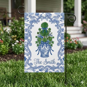 May include: A garden flag featuring a blue and white floral design with a topiary in a vase. The flag has a decorative border and the name "The Smith" in script. The topiary is green with blue ribbons.