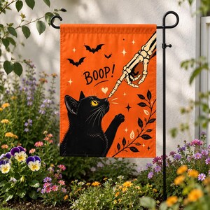 May include: A rectangular orange garden flag featuring a black cat looking up at a skeletal finger touching its nose. The text BOOP is printed in black above the cat, accompanied by a small heart, flying bats, and star-shaped accents.