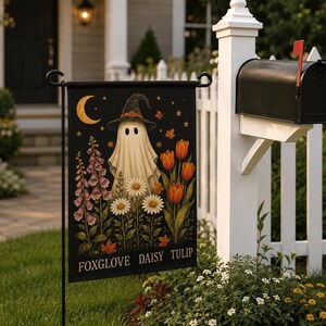 May include: A rectangular black garden flag featuring a central white ghost wearing a witch hat, surrounded by pink foxgloves, white daisies, and orange tulips. A yellow crescent moon and stars appear above. The text FOXGLOVE DAISY TULIP is at the bottom.