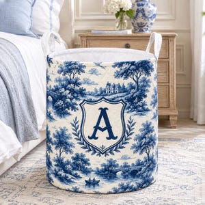May include: A cylindrical quilted laundry hamper featuring a blue and white toile de jouy pattern of landscapes, trees, and a bridge. The front displays a large blue letter A centered within a decorative shield crest with leafy laurel accents.