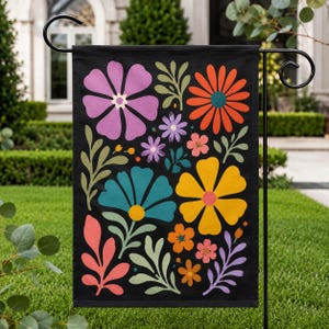 May include: A black garden flag featuring a vibrant floral design. The design includes large, colorful flowers in shades of purple, orange, yellow, and teal, along with green leaves and smaller flowers. The flag is displayed on a black metal stand.