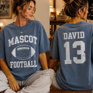 May include: A blue short-sleeve t-shirt featuring a white graphic on the front with the text MASCOT FOOTBALL and an illustration of a football. The back of the shirt displays the name DAVID in white letters above the large white number 13.