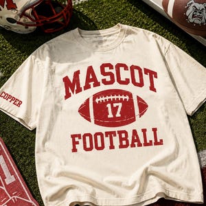 May include: A cream-colored short-sleeve t-shirt featuring red distressed text that reads MASCOT FOOTBALL with a football graphic containing the number 17 in the center. The word COPPER is printed on the right sleeve in red lettering.