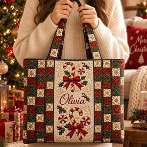 May include: A quilted tote bag featuring a red, green, and white checkered pattern with gold snowflake accents. The center panel displays two candy canes with red bows, holly leaves, and the embroidered name Olivia in red script lettering.