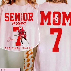 May include: Light pink t-shirt with red text and graphics. The front reads "SENIOR 2026 mom" above a lacrosse player graphic and the name "James". The back displays "MOM of 7". The shirt is a casual style.