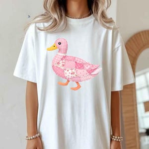May include: White t-shirt featuring a patchwork duck design in shades of pink, with a yellow beak and orange feet. The duck is walking, and the shirt has short sleeves.