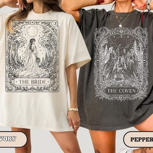 May include: Two oversized t-shirts in ivory and pepper gray. The ivory shirt features a black line art graphic of a bride in a forest with the text THE BRIDE. The pepper shirt displays a white line art graphic of three figures with the text THE COVEN.
